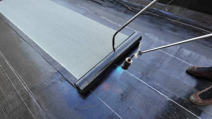 A roofer torches a membrane layer on a flat roof using a tiger torch to seal the asphalt bitumen, ensuring a durable waterproof layer.