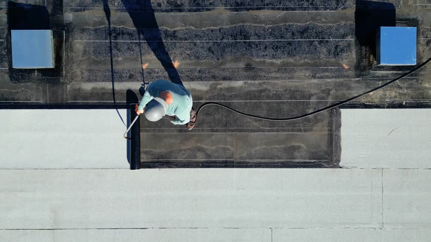 A roofer torches a membrane layer on a flat roof using a tiger torch to seal the asphalt bitumen, ensuring a durable waterproof layer.