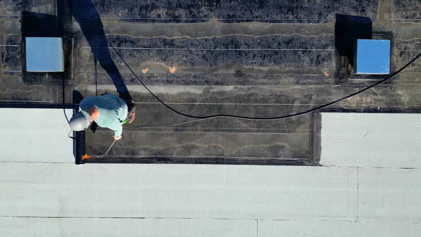 A roofer torches a membrane layer on a flat roof using a tiger torch to seal the asphalt bitumen, ensuring a durable waterproof layer.