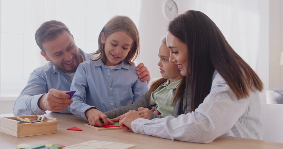 Mother and father playing with wooden geometric figures with little kids at home. Happy family playing logic educational board games with children sitting at desk in living room. 4k video.