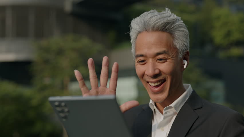 Portrait of joyful Asian businessman with grey hair waves hand on video call via digital tablet outside office. Mature man joins online chat with coworkers on city street - Powered by Shutterstock - Get 15% off with code: PIKWIZARD15
