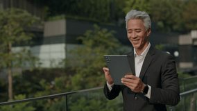 Portrait of middle aged Asian businessman reads report on digital tablet in downtown block. Mature manager smiles and controls market rates via device walking along street - Powered by Shutterstock - Get 15% off with code: PIKWIZARD15