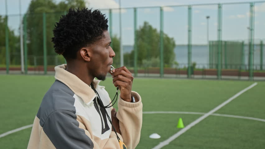 Highly qualified African American coach blowing whistle and turning head to camera, he smiling making confidence impression