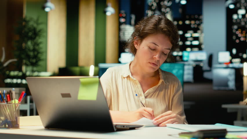 Businesswoman makes mistake in transcribing finance updates on notebook, tearing up the paper sheet and restarting. Discontent impatient worker wanting to finish tasks for the night. Camera B.