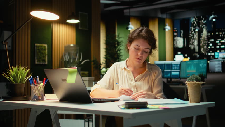 Businesswoman makes mistake in transcribing finance updates on notebook, tearing up the paper sheet and restarting. Discontent impatient worker wanting to finish tasks for the night. Camera A.