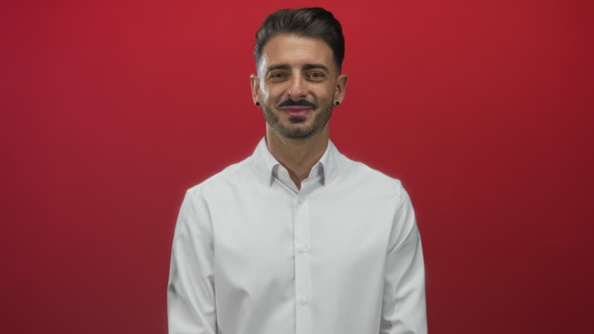 Hispanic man face winking with toothy smile and confident gaze wearing white shirt in red studio; playful charm cheerful.