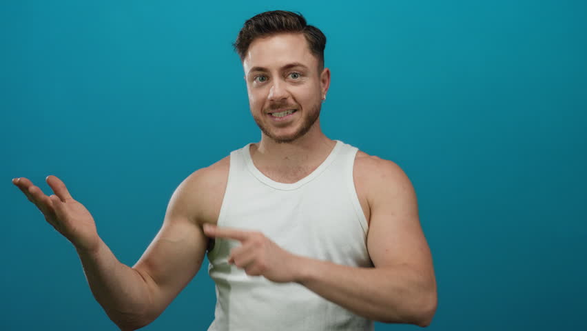 Young man with a beard in a white tank top smiling and pointing to an open hand against a solid blue background, conveying a positive and inviting presentation gesture. - Powered by Shutterstock - Get 15% off with code: PIKWIZARD15