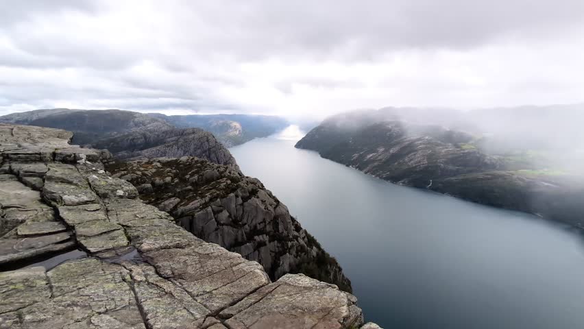 Breath-taking aerial view from Norway’s iconic Preikestolen (Pulpit Rock), overlooking a dramatic fjord surrounded by steep mountains and misty clouds on a rainy day. Scenic 4K horizontal footage.