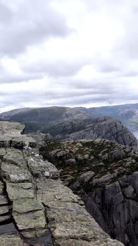 Dramatic POV shot from the edge of Preikestolen (Pulpit Rock) in Norway, looking down over the majestic fjord surrounded by towering cliffs and low-hanging clouds on a rainy day. Vertical 4K footage.