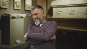 Man smiling with arms crossed and one hand touching ear amid stone sculptures in an art gallery building, casual shirt and relaxed pose; amusement. - Powered by Shutterstock - Get 15% off with code: PIKWIZARD15
