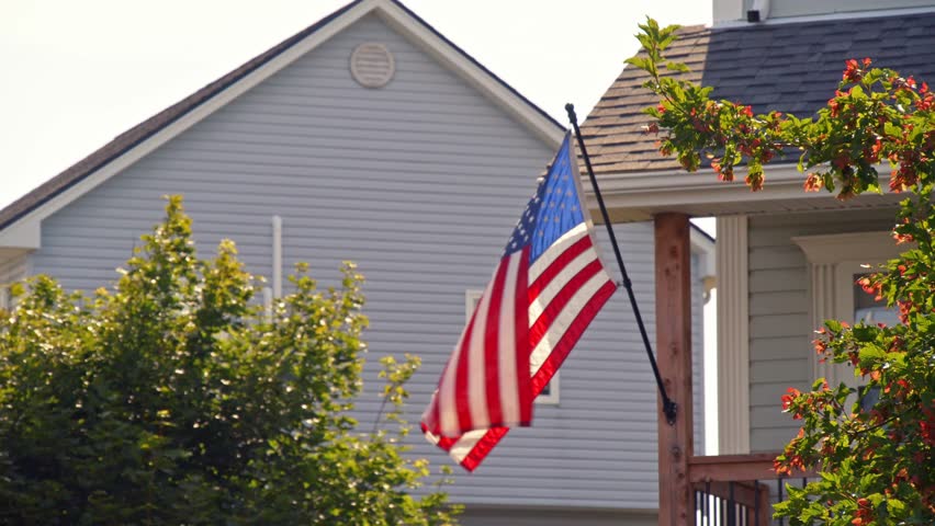 Traditional Flag On A Pole Next To A Rural American House. Trees, Field, And Blue Sky Create A Peaceful, Patriotic Scene On A Warm Day In The United States.American Flag By House In Countryside