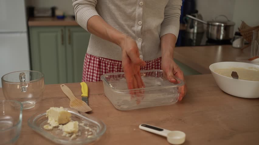 Young woman in a checkered apron preparing a delicious homemade pie, first greasing the glass baking dish with butter and then carefully pouring the fresh batter from a white bowl