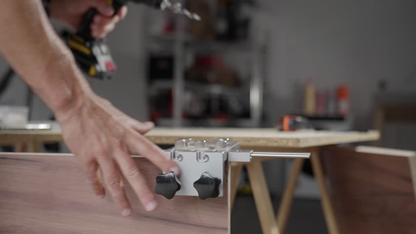Close-up of carpenter drilling hole in wooden panel using drill and metal jig tool for precision. Man working in home workshop. Concept of woodworking, furniture making and DIY home projects