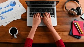 Woman types on laptop at wooden desk. Coffee cup, headphones, notebook, pen and charts are also visible. Work from Home or Freelancer concept. - Powered by Shutterstock - Get 15% off with code: PIKWIZARD15