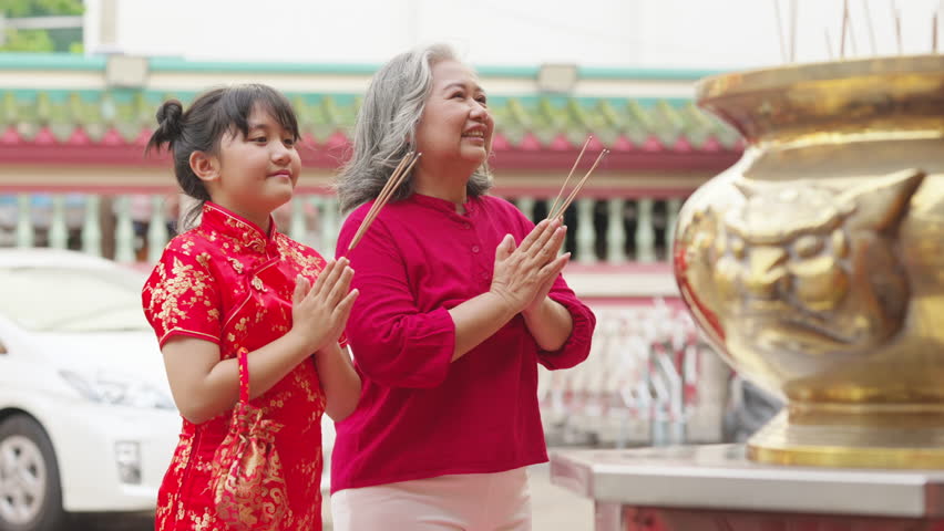 Chinese lunar new year festive holiday event celebration. Happy Asian family grandmother and grandchild girl praying a wish for health, wealth and luck with incense and red candle at Chinese shrine.