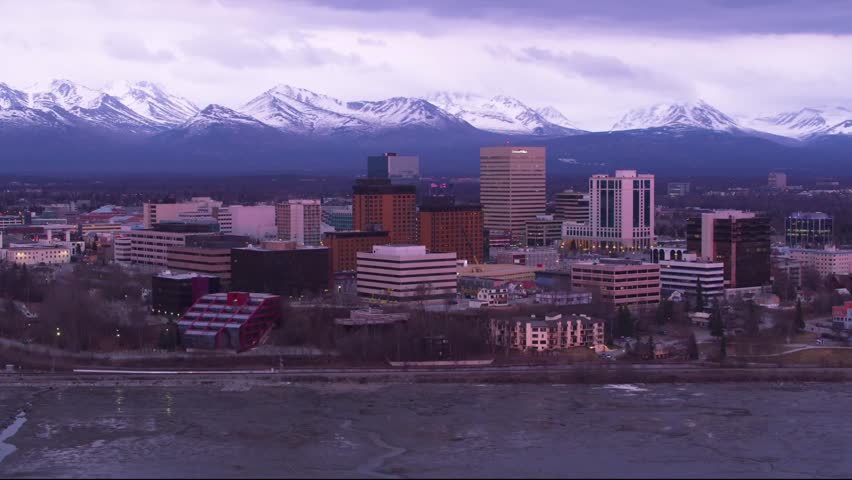 The shot is taken from an elevated perspective, capturing the dense urban core of Downtown Anchorage. Looming dramatically behind the city are the magnificent Chugach Mountains, 
