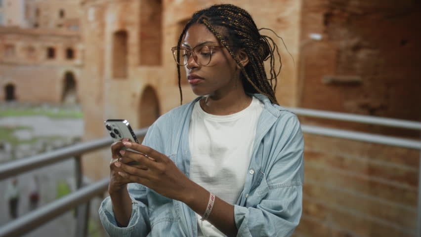 African american woman holding smartphone at roman ruins building, hands visible scrolling screen while glancing aside; contemplation curiosity.