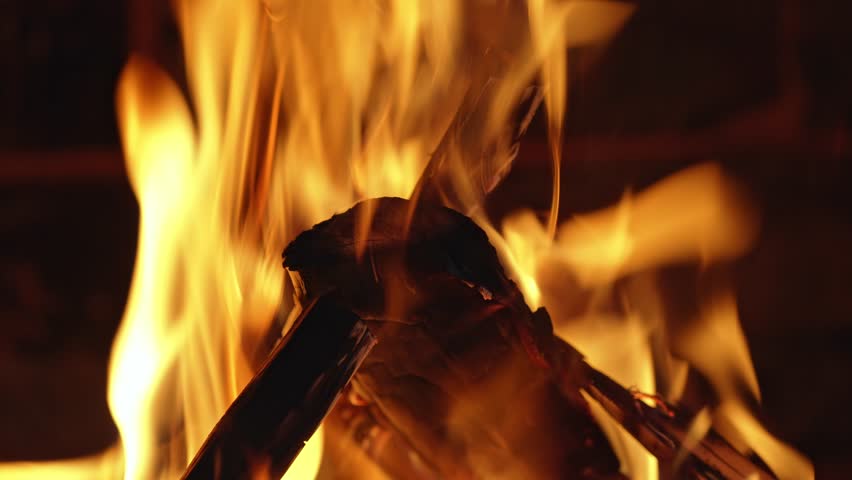 Bright orange and yellow flames dancing in slow motion around burning logs in a campfire. The cozy and warm fire provides a mesmerizing background of heat, light, and beautiful combustion