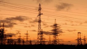 Several electricity pylons stand silhouetted against a warm orange sunset sky. Many power lines stretch between the industrial high voltage transmission towers. - Powered by Shutterstock - Get 15% off with code: PIKWIZARD15
