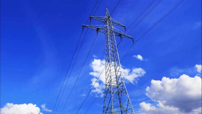 Tall transmission tower stands against vivid blue sky. Power lines extend from structure. Fluffy white clouds drift in background. Industrial landscape.