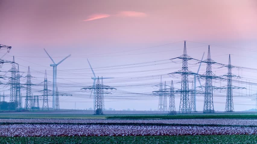Wind turbines rotate near power line towers across fields. Light fades in background. Power distribution and renewable energy coexist in rural environment.