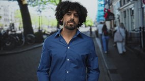 Man with curly hair shrugs with open hands on a city street, blue shirt, visible palm, surrounded by blurred pedestrians and bicycles; confusion. - Powered by Shutterstock - Get 15% off with code: PIKWIZARD15