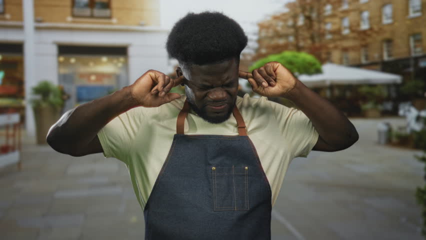 Man plugging fingers into ears on street outside cafe wearing denim apron, eyes closed and mouth open; annoyance urban noise.