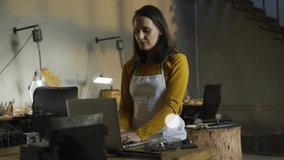 Mid-adult woman typing on laptop in studio visualizing tech network with smartphone and music icons. Workshop, artisan, connectivity, digital, crafting, innovation, workspace - Powered by Shutterstock - Get 15% off with code: PIKWIZARD15