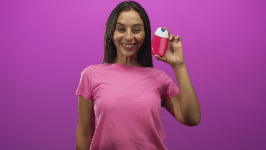 Young woman holds pink lotion bottle and beckons with outstretched hand in magenta studio; playful skincare demo.
