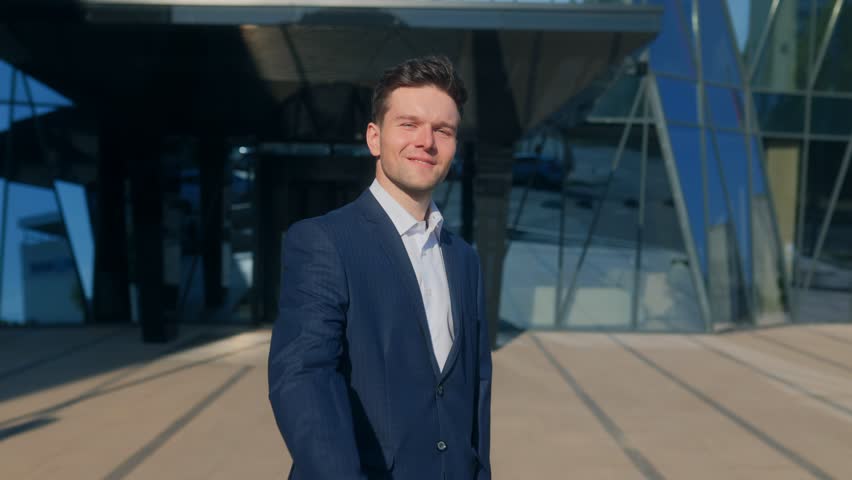 A charismatic young man in a suit makes a follow me hand gesture, smiling confidently in front of a modern glass office building, inviting others with charm and energy.