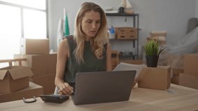 Young woman with blonde hair using a calculator while working on a laptop in her new home surrounded by moving boxes in a living room with interior decor and a potted plant. - Powered by Shutterstock - Get 15% off with code: PIKWIZARD15