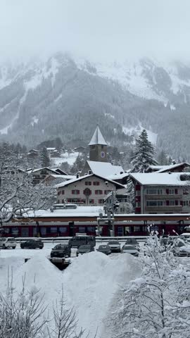 Red passenger train pulling into a station in a picturesque alpine village during a heavy snowfall
