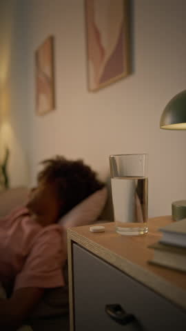 Medium vertical shot of young African American woman lying in bed, getting up, dropping soluble painkiller medicine tablet into water and drinking, while suffering from migraine headache