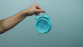 Man's hand holding a turquoise alarm clock against a blue background, with a pointing gesture towards the clock, suggesting urgency or time management. - Powered by Shutterstock - Get 15% off with code: PIKWIZARD15