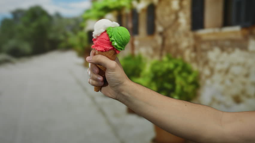 Man holding colorful ice cream cone outdoors with blurred city street background, showcasing summer refreshment and urban leisure.