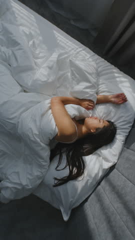 In Morning, Young Girl Sleeps in Cozy Bed in Stylish Bedroom. Asian Woman in White Bedding, her Dark Hair Lies Beautifully on Pillow. Concept Healthy Sleep, Beautiful Morning Routine. Vertical Shot.