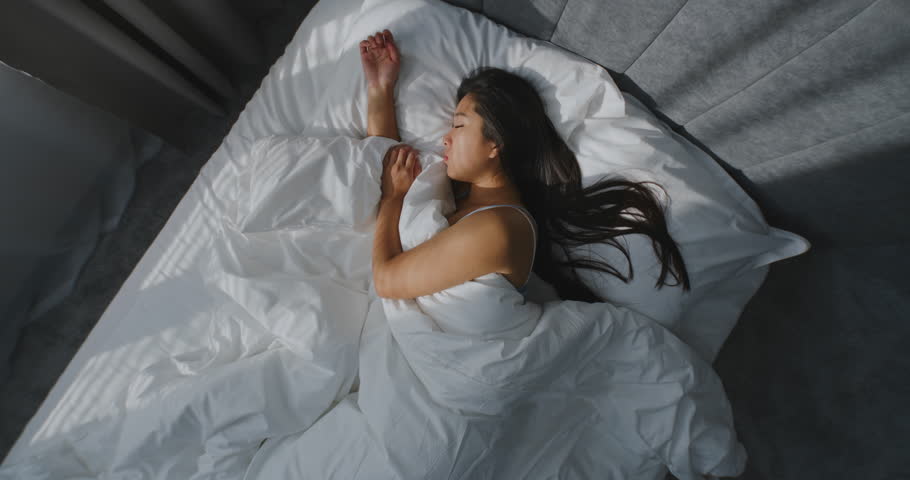 In Morning, Young Girl Sleeps in Cozy Bed in Stylish Bedroom. Asian Woman in White Bedding, her Dark Hair Lies Beautifully on Pillow. Concept of Healthy Sleep, Rest, and Beautiful Morning Routine.
