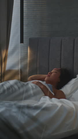 Asian Woman in Nightwear Sits on Bed By Window in Soft Morning Light. Girl Wakes Up in Bed, in Dreamy and Pensive Mood. Peaceful Moment of Calm Reflection, Cozy Lifestyle, Home Comfort. Vertical Shot.