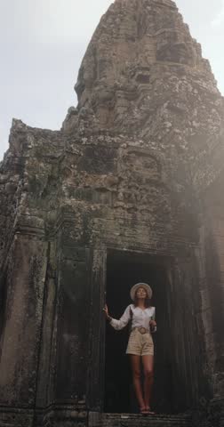 Cinematic vertical shot of a young woman in hat and shorts standing at the doorway of Bayon Temple in Angkor Thom, Cambodia. High quality stock video for travel, tourism, culture, and lifestyle promo.