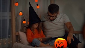 Father and little daughter in witch hat watch horror film on tablet in dark with orange pumpkin lantern. People use tablet. Halloween - Powered by Shutterstock - Get 15% off with code: PIKWIZARD15