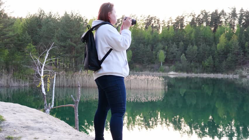 A woman stands by a calm lake, taking pictures of the beautiful scenery. The sun rises, illuminating the lush trees and creating stunning reflections on the water