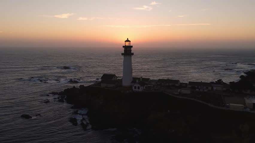 The sun sets over the Pacific Ocean near Pigeon Point Lighthouse, casting warm hues on the sky and rippling waves with rocky outcrops in view.