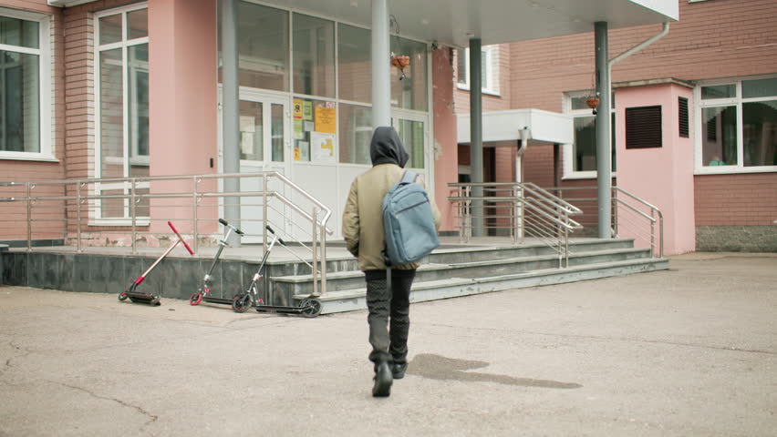rear view student with backpack walking toward school entrance on overcast day, scooters parked by steps, urban campus exterior, quiet morning arrival, education theme, safe environment