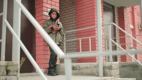 Teen in hooded jacket leans on building wall outdoors operating smartphone while holding brown book, focused on screen, standing near stair railing against red brick background - Powered by Shutterstock - Get 15% off with code: PIKWIZARD15