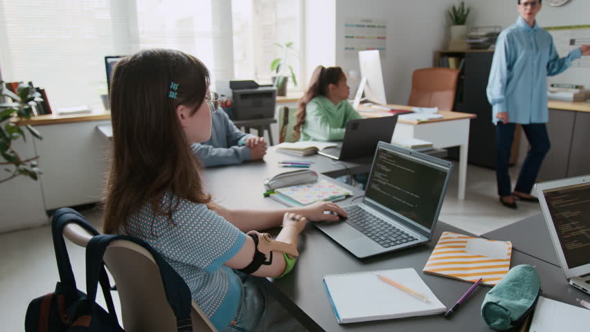 High angle handheld zoom in shot of young girl with prosthetic arm attending programming lesson in modern school classroom