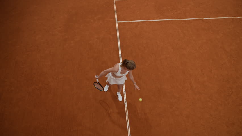 High angle shot of focused young adult woman in white tennis set bouncing ball against clay court surface then serving strong overhead shot during sports match, slow motion