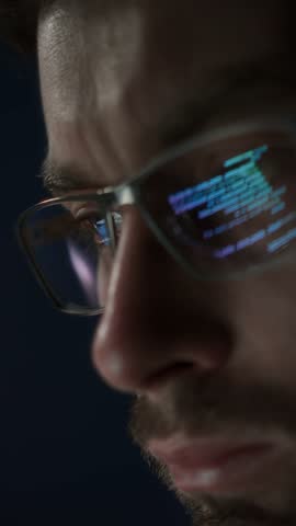 Close up of concentrated programmer in glasses works late at night, lines of code reflecting in his eyeglasses. Software engineer is engaged in coding, data science, or AI development. Vertical video