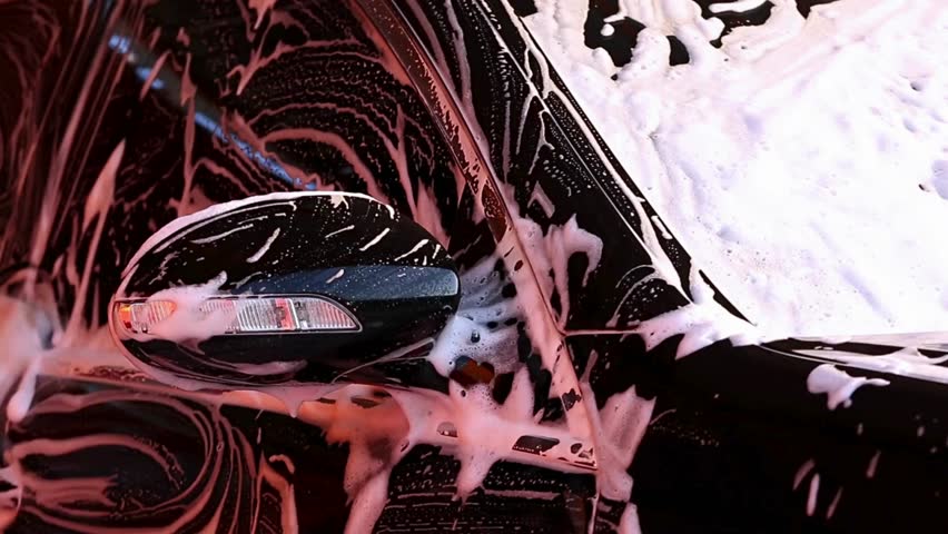 Hands apply soap suds to a vehicle surface during the car washing process. The focus is on foam coverage and cleaning technique for a spotless finish.