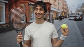 Young man wearing wristbands and a white tee holds a tennis racket and grips a neon ball on a bustling city street; enthusiasm. - Powered by Shutterstock - Get 15% off with code: PIKWIZARD15
