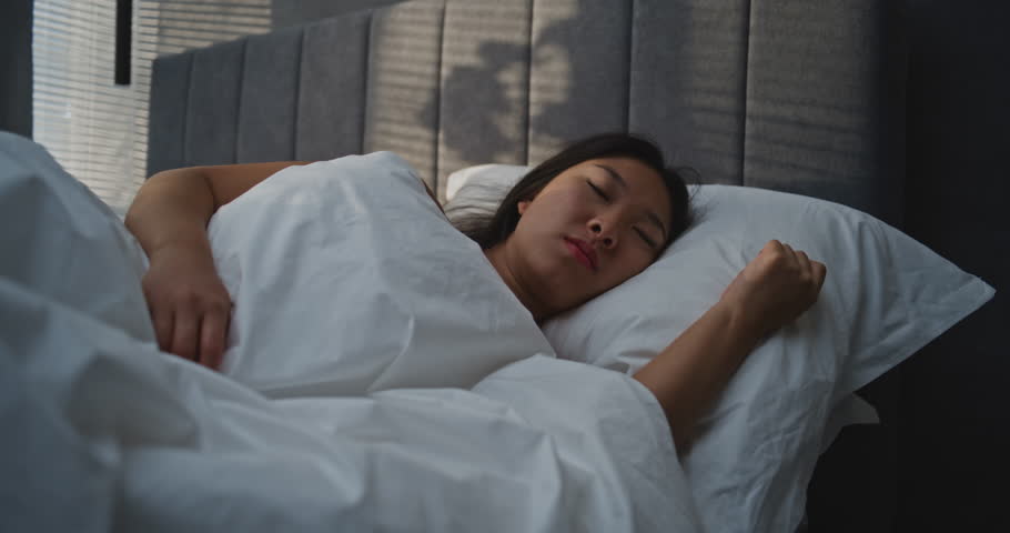 Beautiful Asian Girl Wakes Up in Bedroom, Squinting in Bright Sunlight. Young Woman Sits Up in Bed, Stretches, and Smiles at Bright Sun. Concept of Healthy Sleep, Beautiful Morning Routine. Handheld.